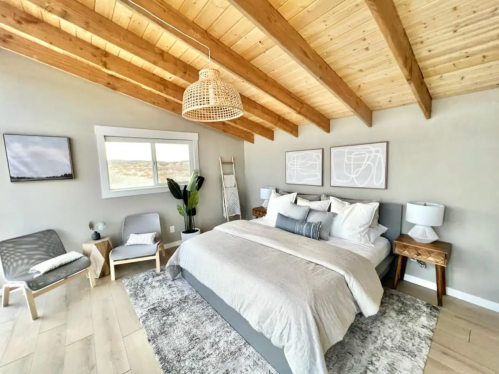 Bright, modern bedroom with a large bed, wooden beams, and minimalist decor, featuring a plant and cozy seating.